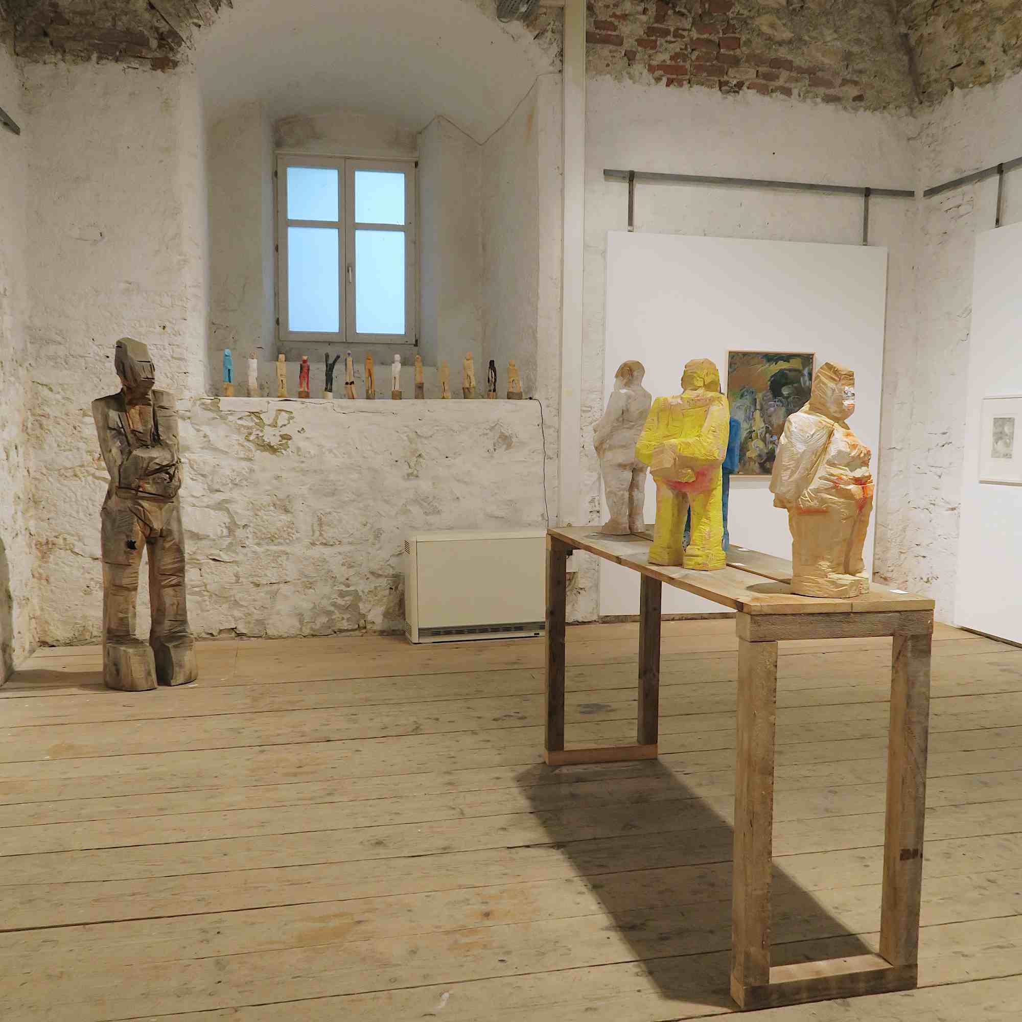 Blick in den Nebenraum der Kunstausstellung 'WAS BLEIBT - WAS ZÄHLT' in Ingolstadt mit kleinen Holzskulpturen und der lebensgroßen Holzskulptur 'Heinz'